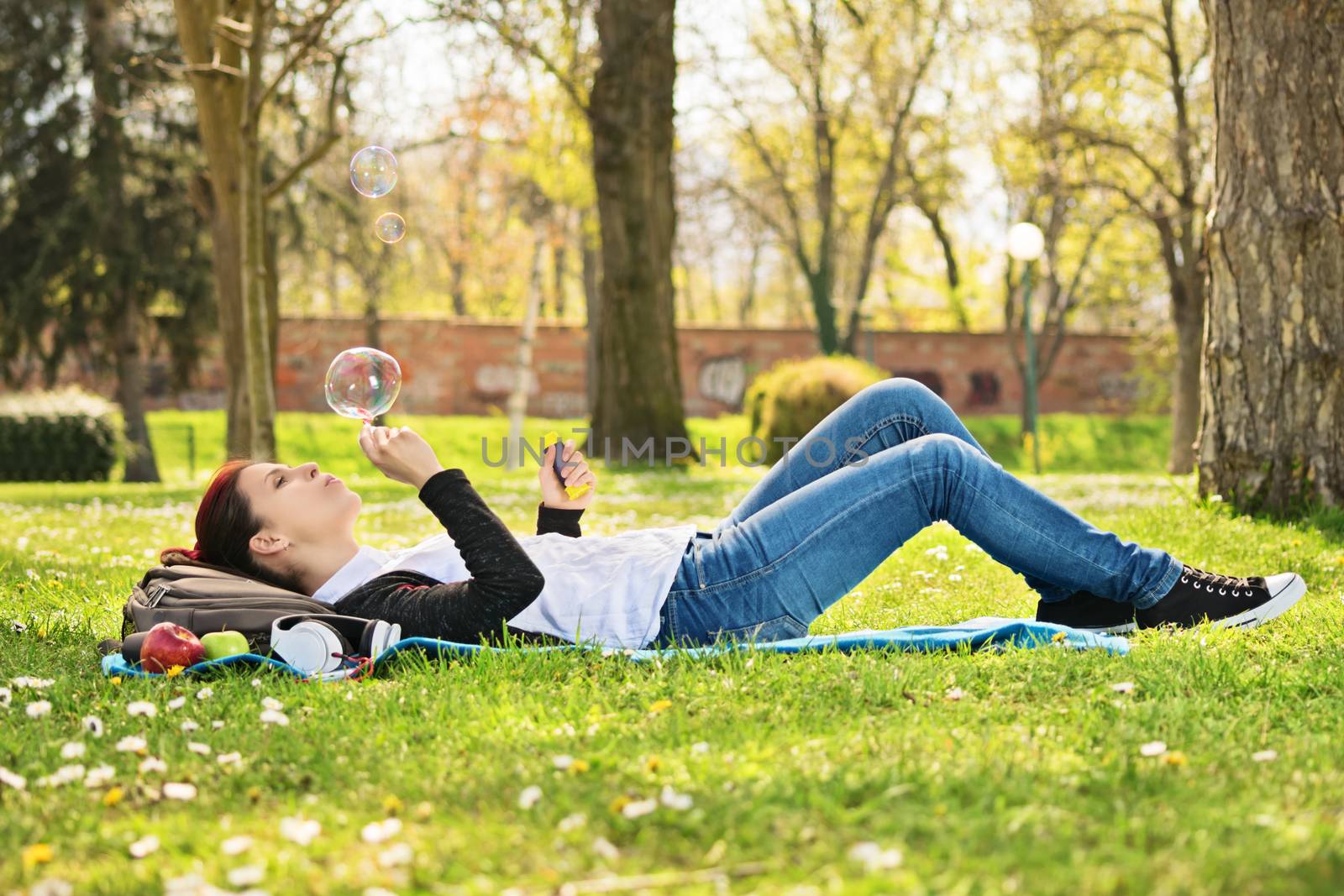 Daydreaming in the park. Beautiful young student girl lying on a meadow blowing soap bubbles, taking a break from studying.