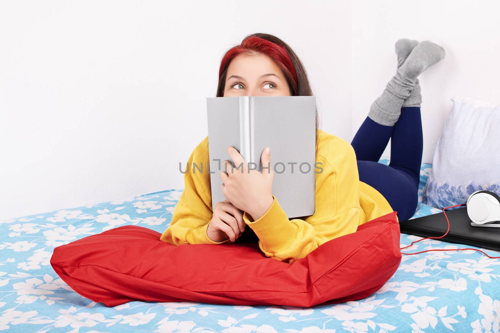 Young beautiful girl lying on the bed in a bedroom, next to a laptop and headphones, holding a book over her face, trying to study but instead she is daydreaming of being somewhere else, isolated on a white background.