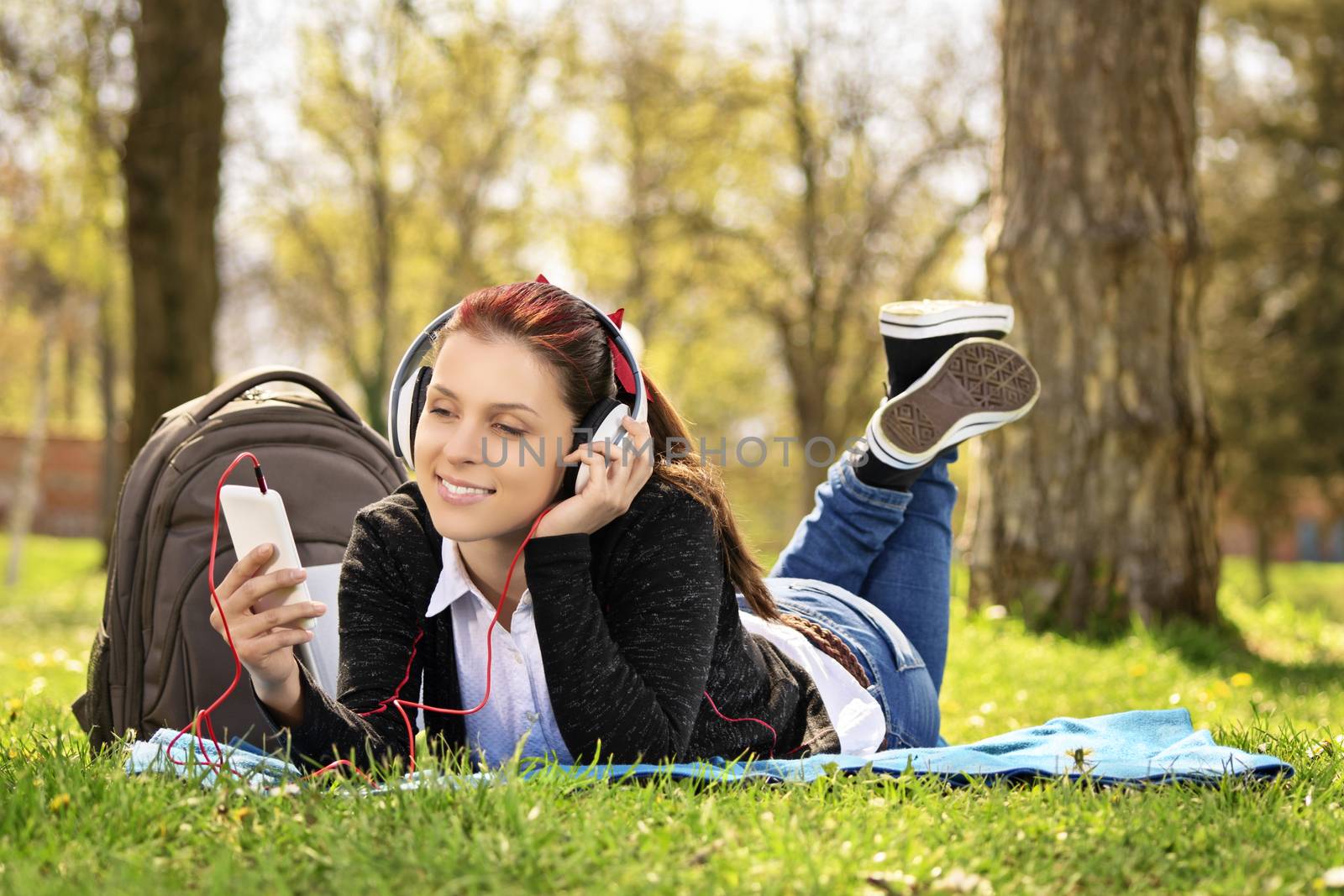 Young beautiful girl lying on a blanket on the grass in the park, listening to music and looking at the phone.