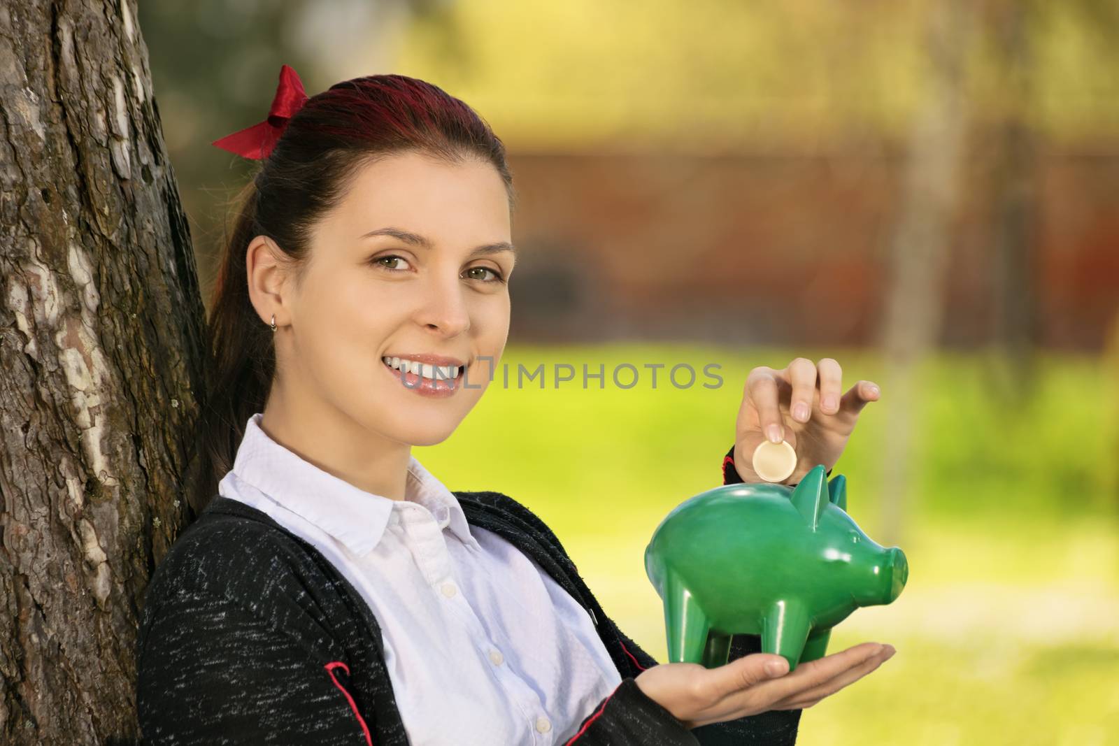 Saving during sunny days so when the rainy ones come. Beautiful young female student leaning on a tree in the park, putting a coin in a piggy bank.