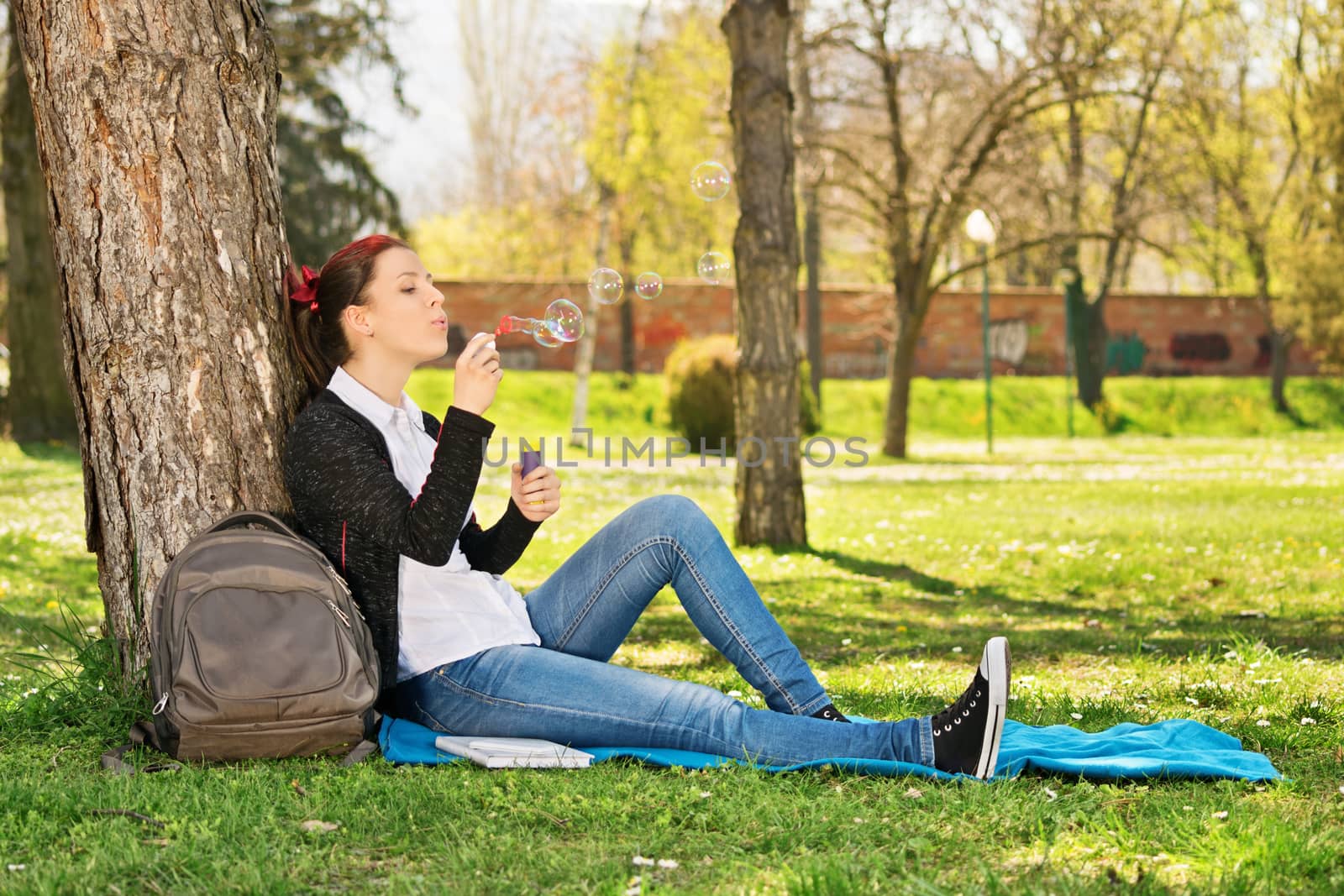 Blowing soap bubbles in the park. Beautiful young student girl leaning on a tree in a meadow blowing soap bubbles, taking a break from studying.