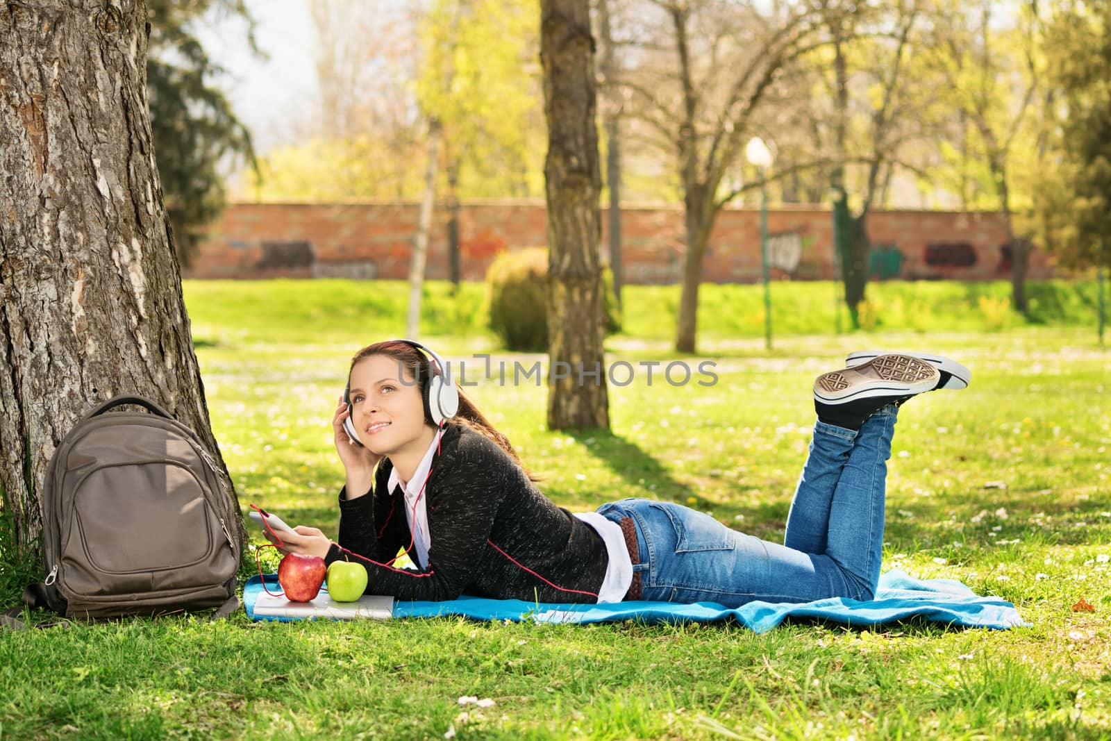 Daydreaming in the park listening music. Beautiful female student, lying on a blanket on the grass in a park listening to music. Student taking a break from studying.