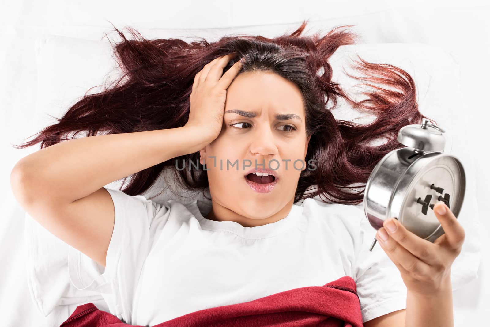 I overslept again! A girl lying in bed and holding her head looking at the alarm clock realizing that she is late, isolated on white background.