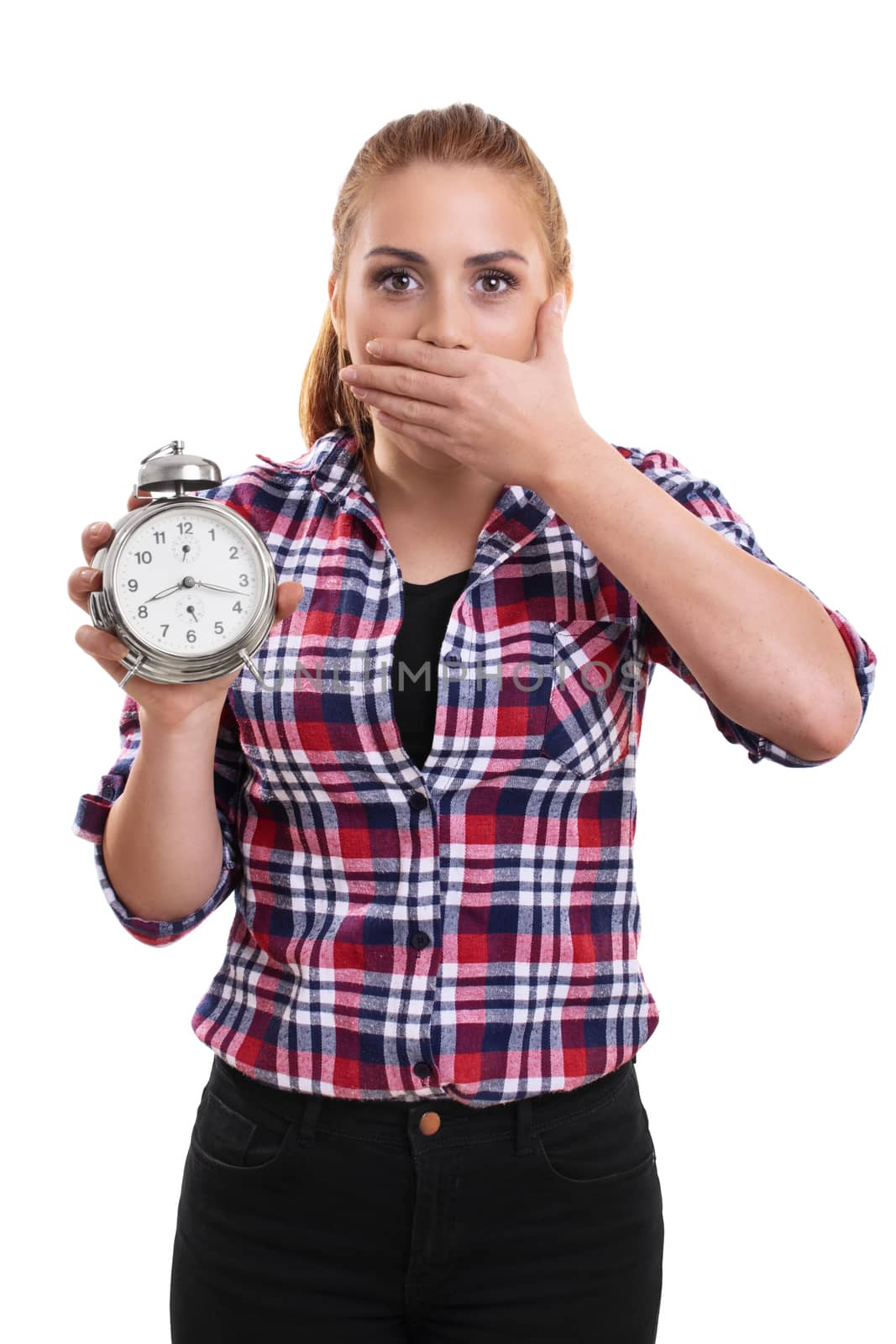Portrait of a young beautiful woman dressed in plaid shirt holding an old school alarm clock, shocked and surprised to see what's the time. Isolated on a white background.