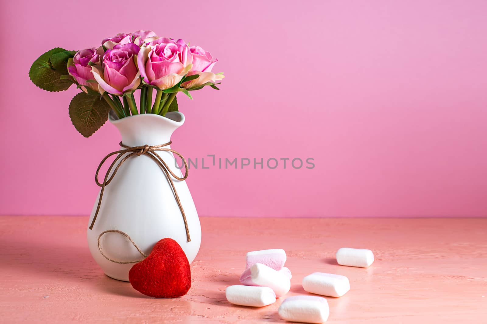 Small white and pink marshmallows are scattered on a pale pink background next to a vase of roses. Place for text.