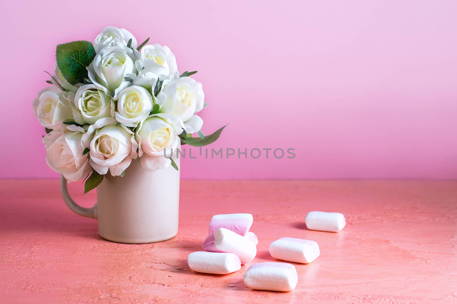 Small white and pink marshmallows are scattered on a pale pink background next to a vase of roses. Place for text.