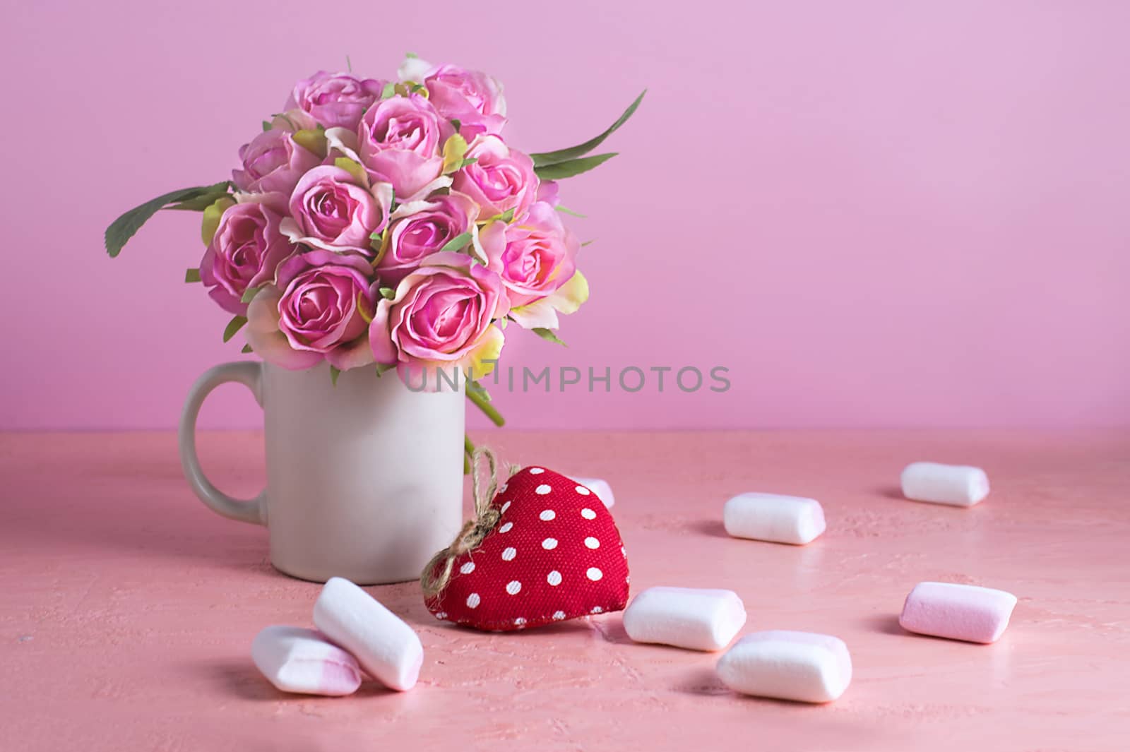 Small white and pink marshmallows are scattered on a pale pink background next to a vase of roses. Place for text.