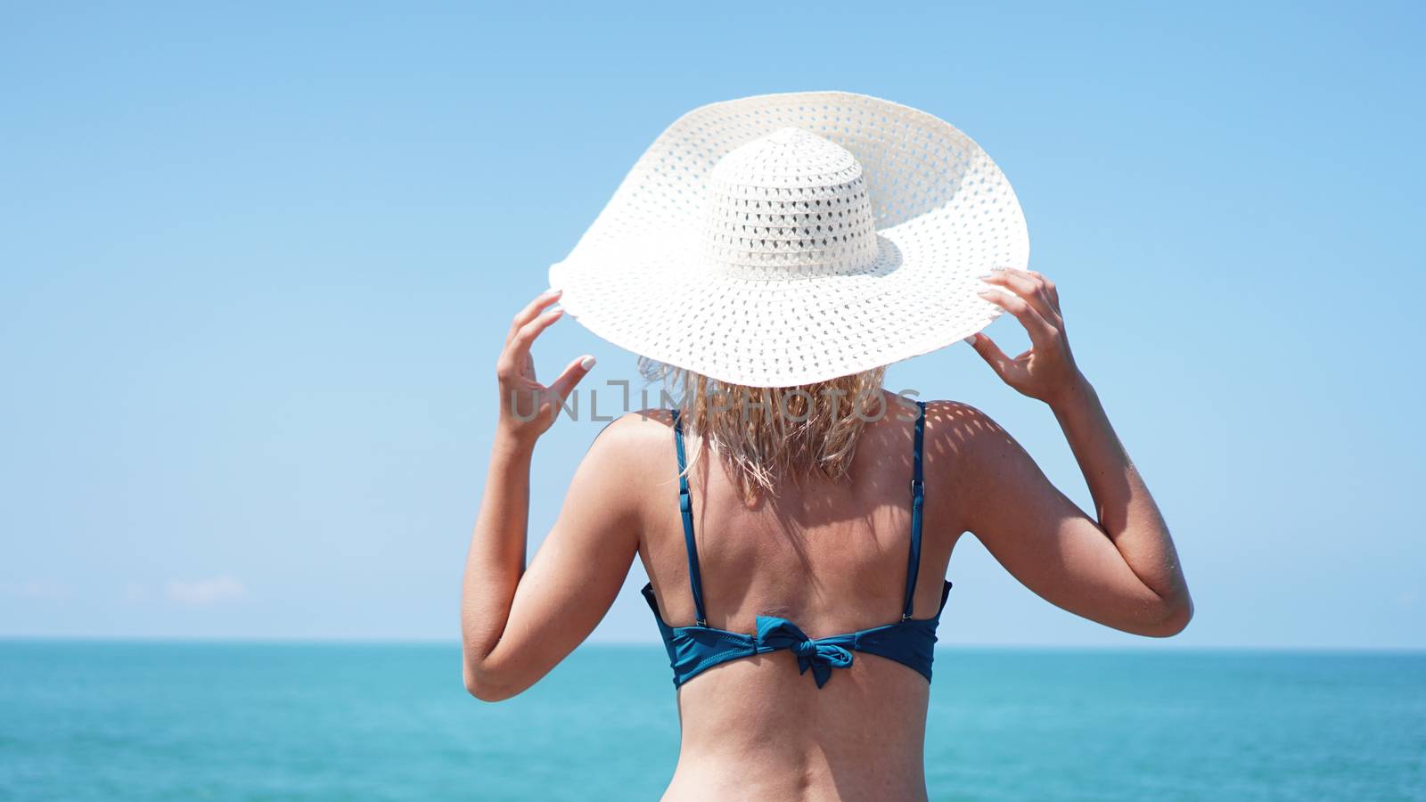 Young woman standing on sand near sea and holding a white hat