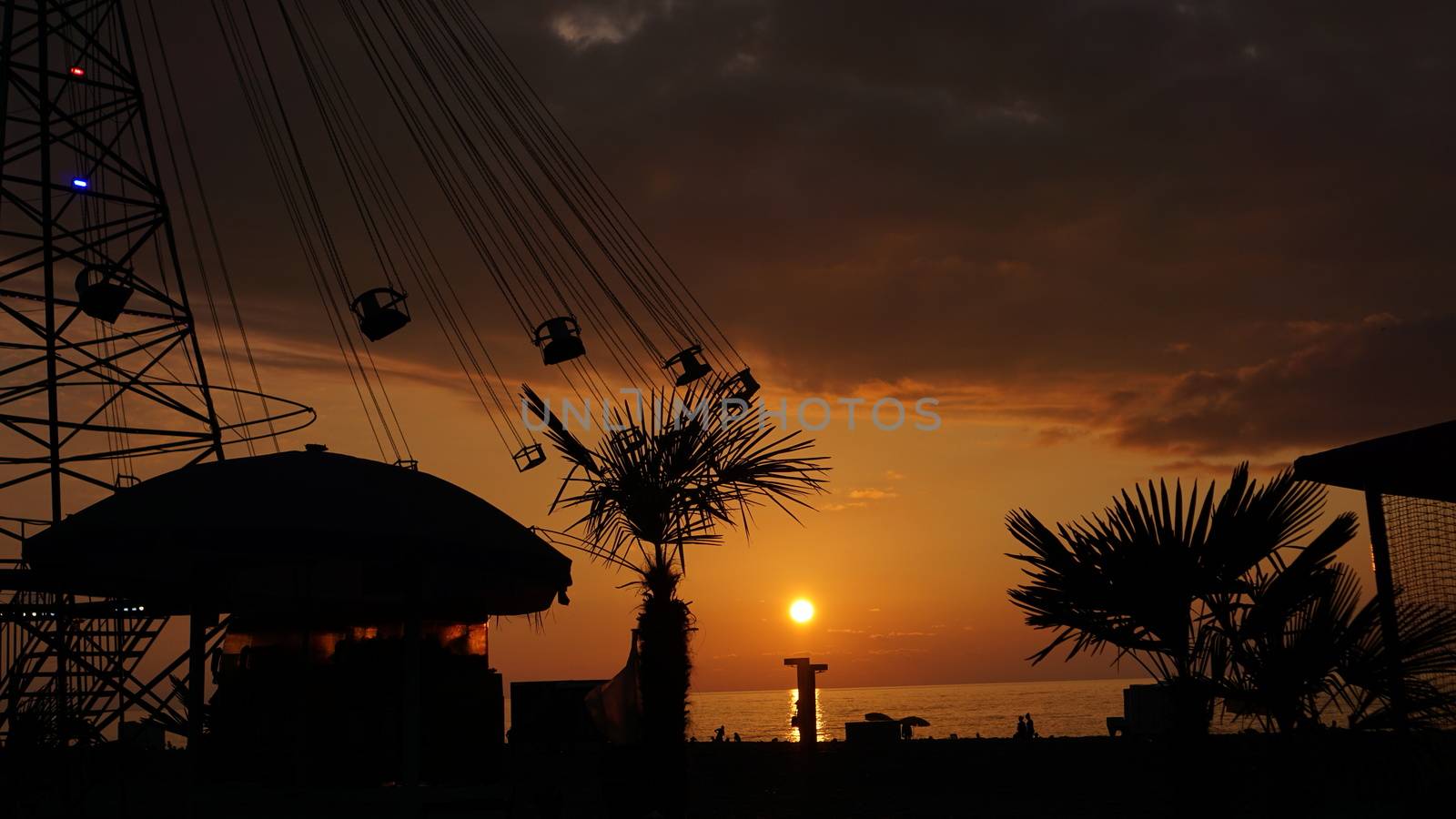 Swinging carousel roundabout chain ride at sunset. Entertainment on the beach, silhouettes of palm trees on a background of sea sunset