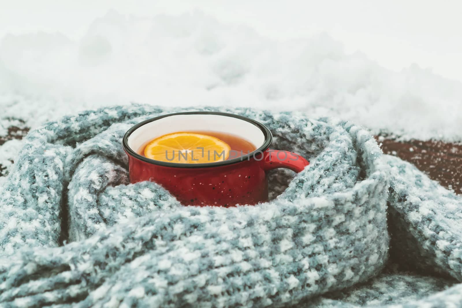 Red enameled cup of hot tea with lemon wrapped in a knitted scarf on a snowy wooden table.