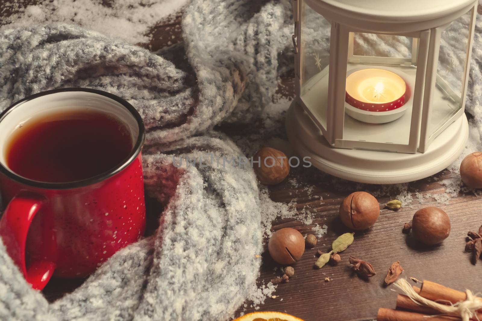 Lantern with a burning candle, spices and a red mug with hot coffee on a snowy wooden table.