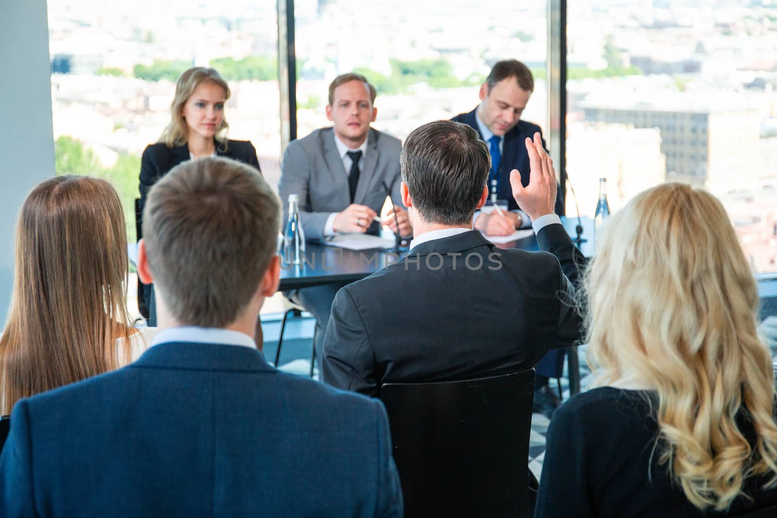 Audience of business people sitting at training room and talking, speakers sitting at the table with documents, microphones and water