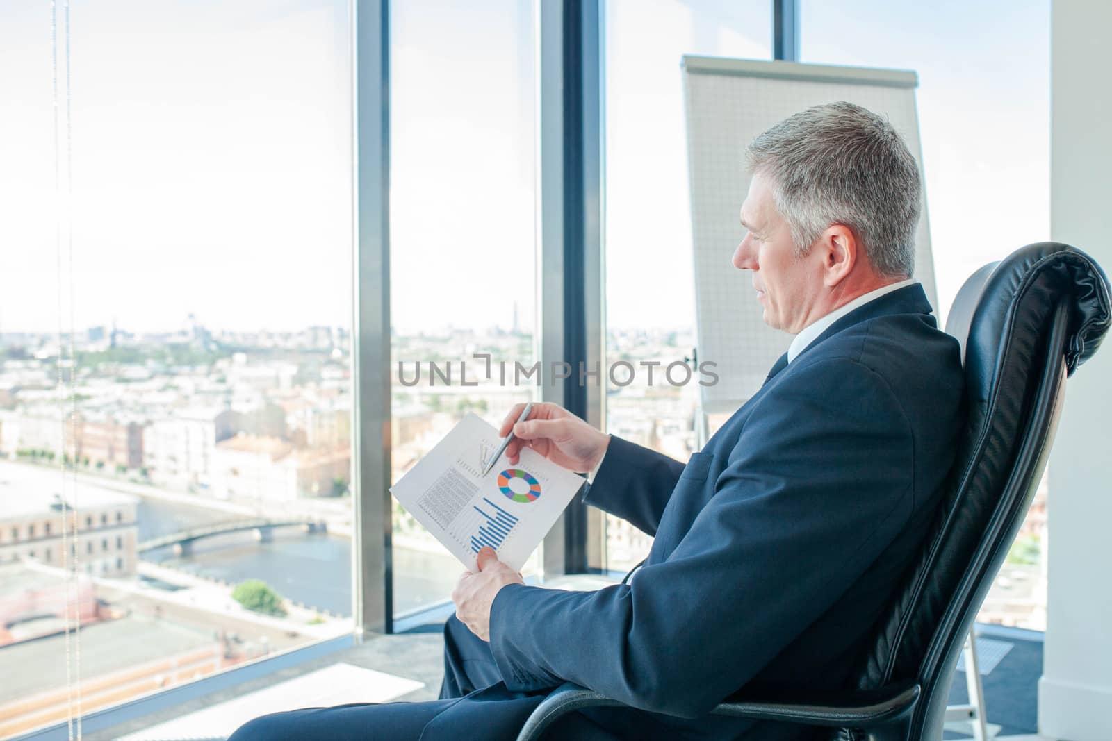 Mature businessman sitting alone in office with large panoramic windows with view at city and looking at diagrams and financial reports