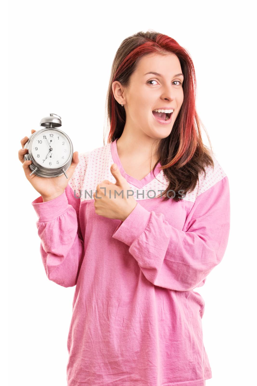 When timing is right. Smiling young girl in pink pajamas holding an alarm clock and giving thumbs up, isolated on white background.