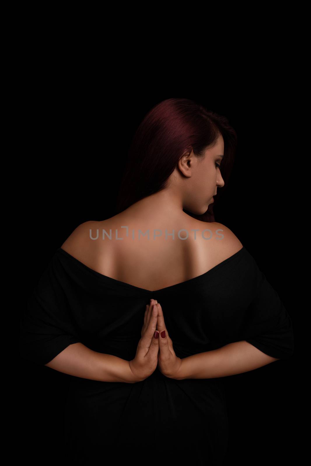 Low key portrait of a young girl in black dress with her hands behind her back, practicing yoga isolated on black background.