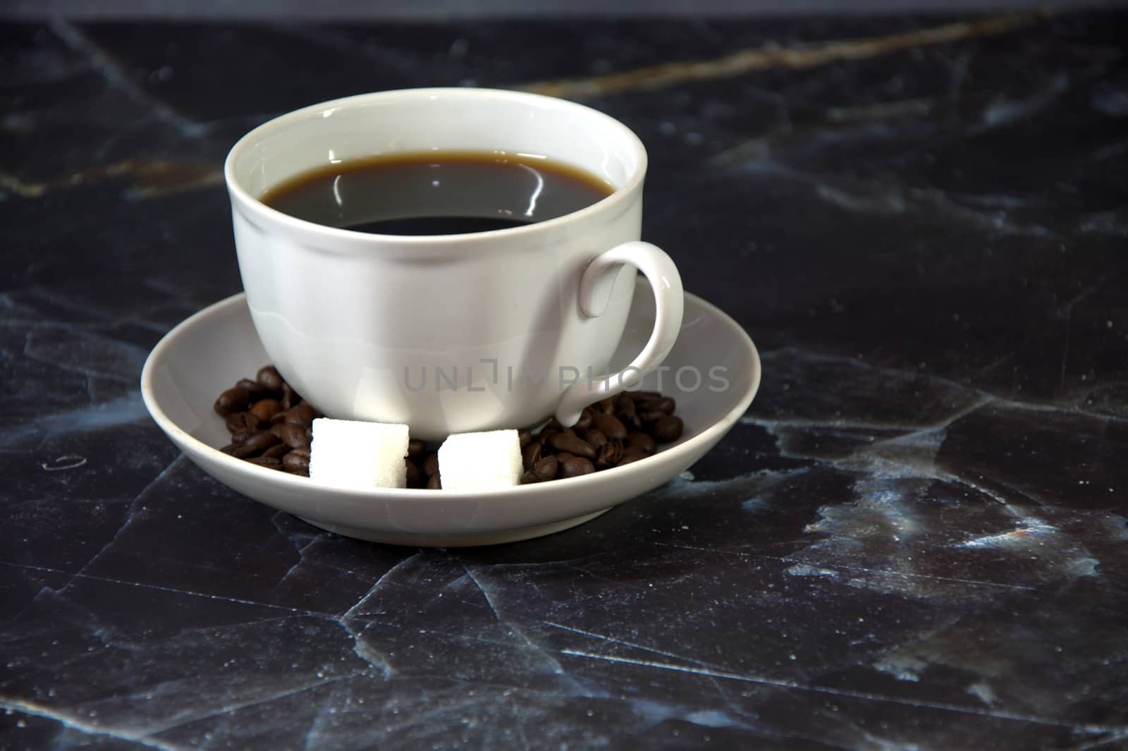 A white ceramic cup of black coffee on a saucer with two cubes of sugar and sprinkled coffee beans. Close-up.