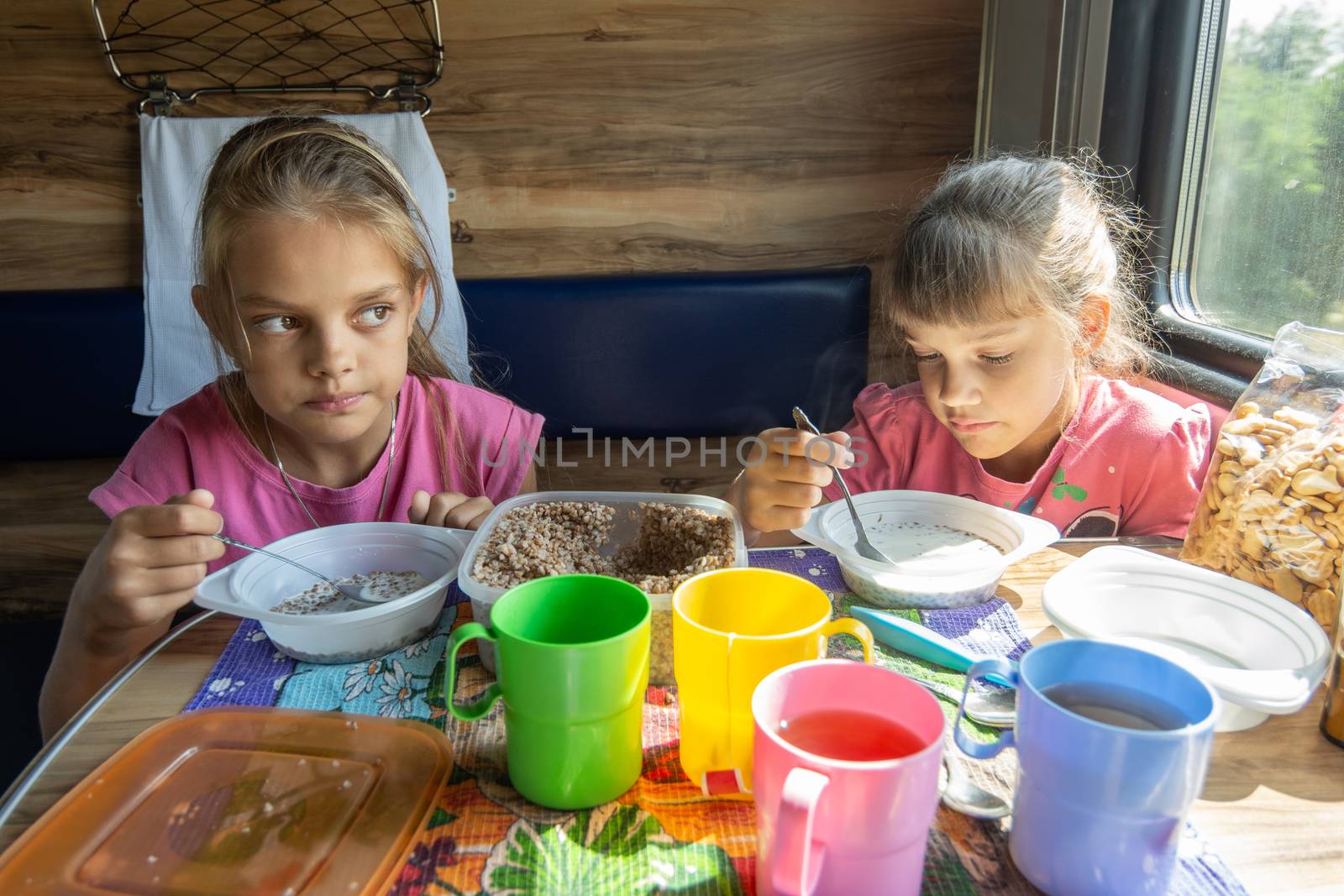 Two girls eat buckwheat with milk from plastic disposable tableware on a train by Madhourse