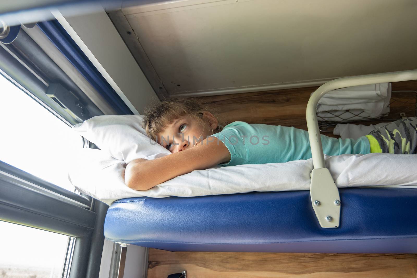 An eight-year-old girl lies on the top shelf of a reserved seat car and looks tiredly out the window