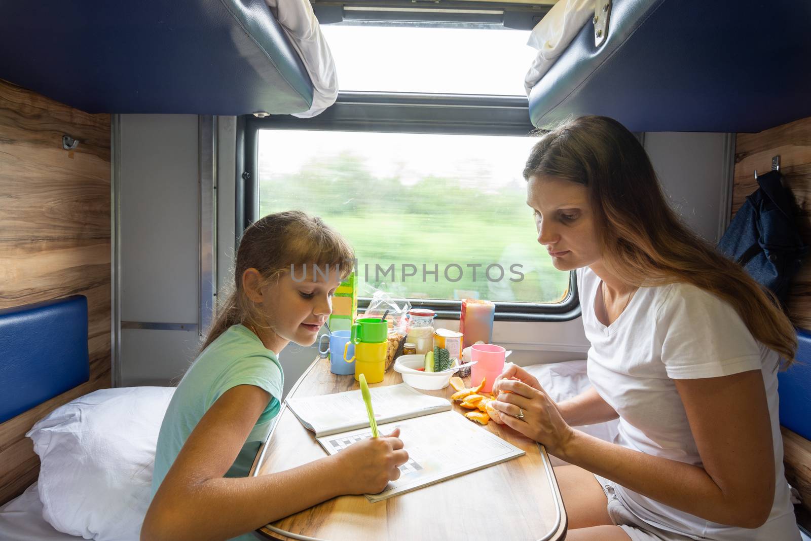 Mom and daughter sit at the table in the train and solve the puzzle in the children's magazine