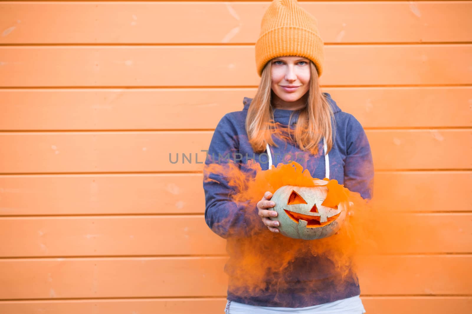 Woman in casual clothes and hat holding Halloween pumpkin over orange wall background with copy space for text