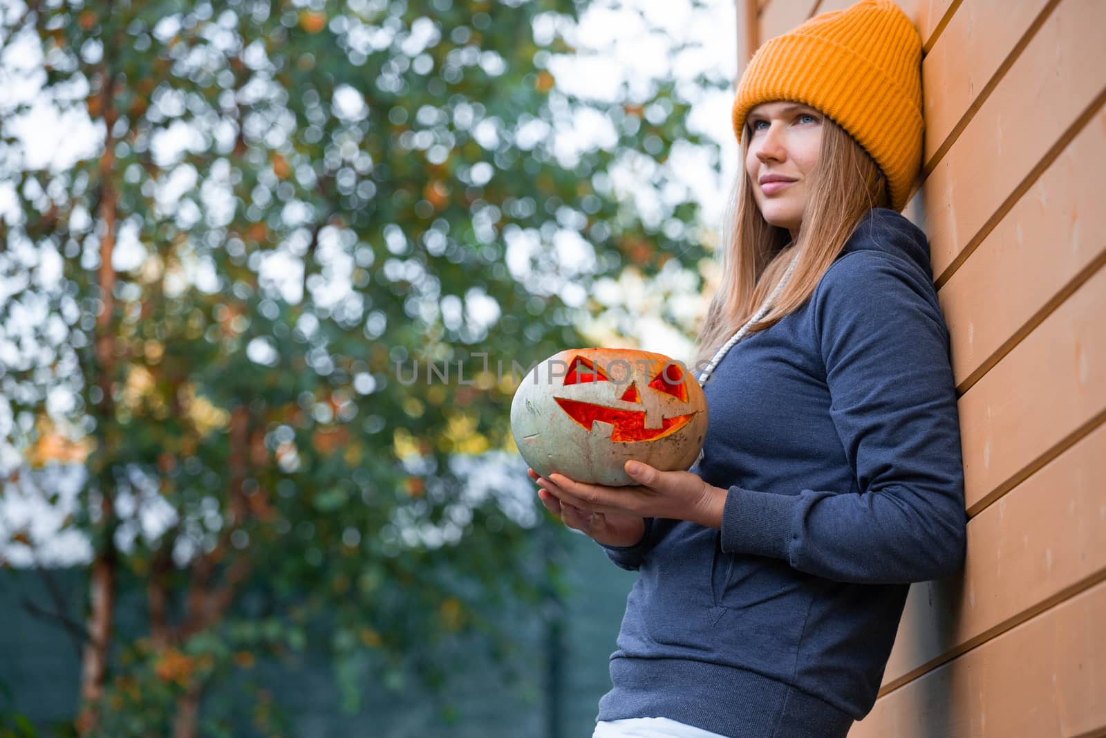 Woman in casual clothes and hat holding Halloween pumpkin over orange wall background with copy space for text