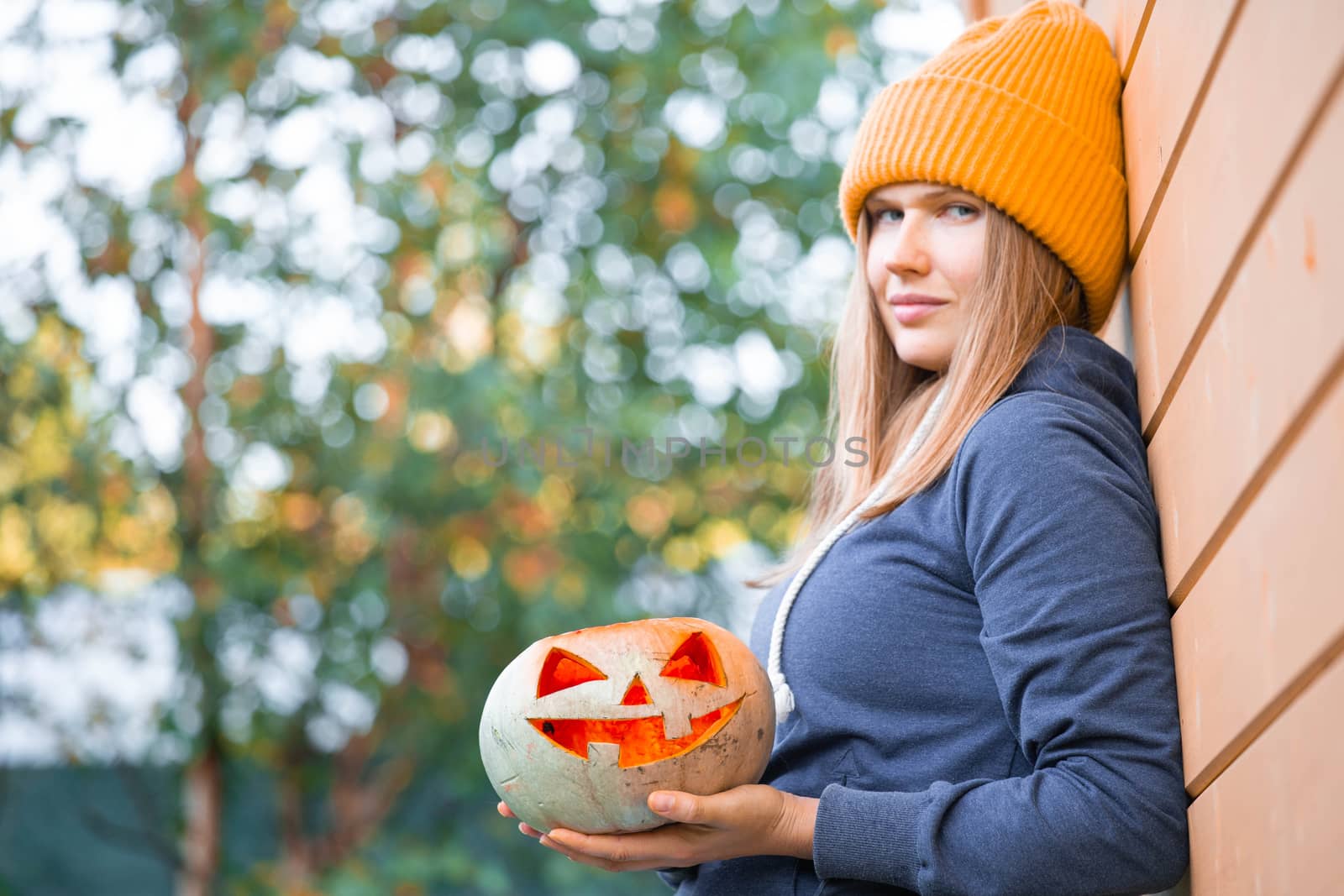 Woman in casual clothes and hat holding Halloween pumpkin over orange wall background with copy space for text