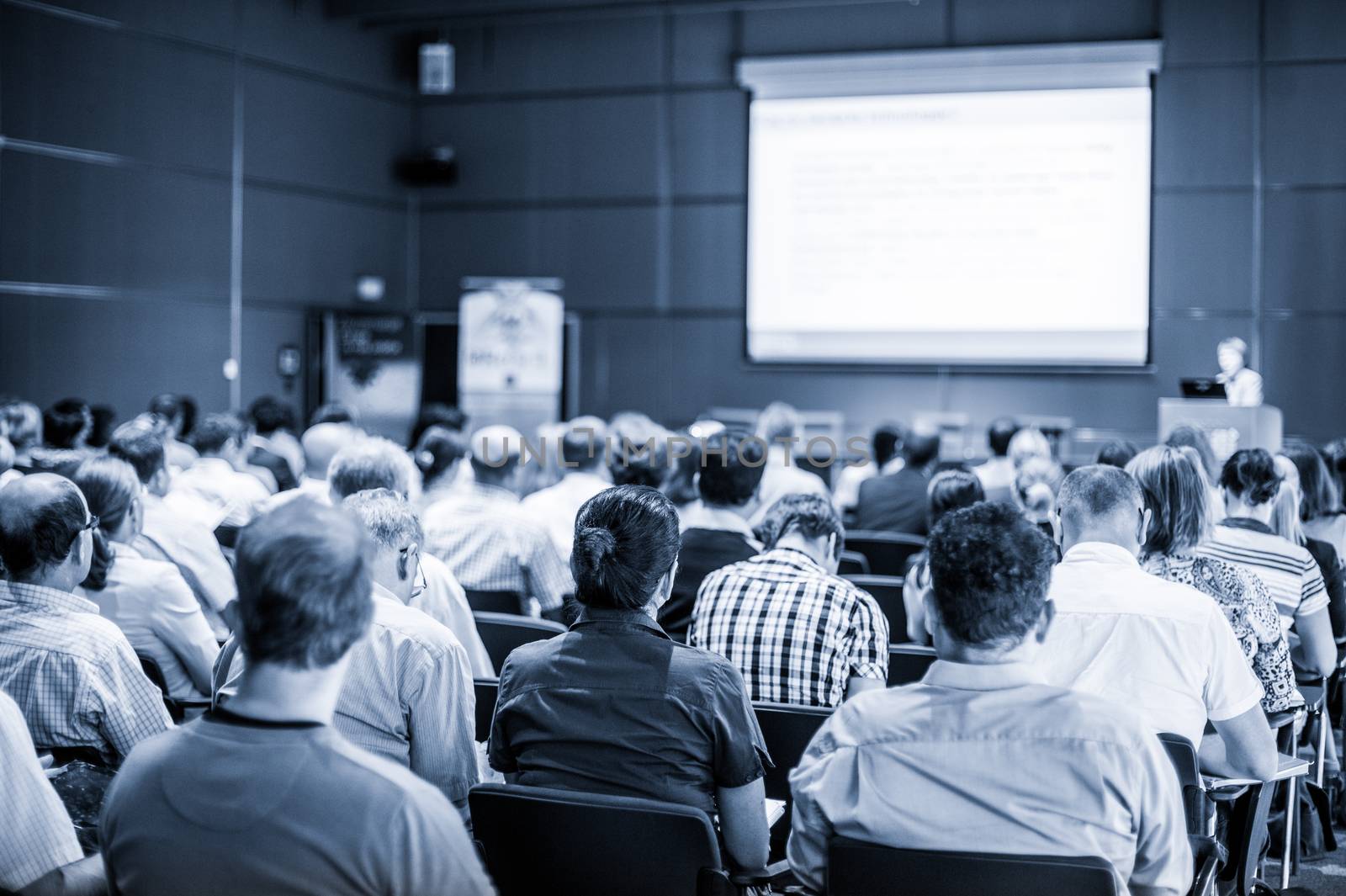 Speaker giving talk at business event. Audience at the conference hall. Business and Entrepreneurship concept. Focus on unrecognizable people in audience. Blue toned grayscale image.