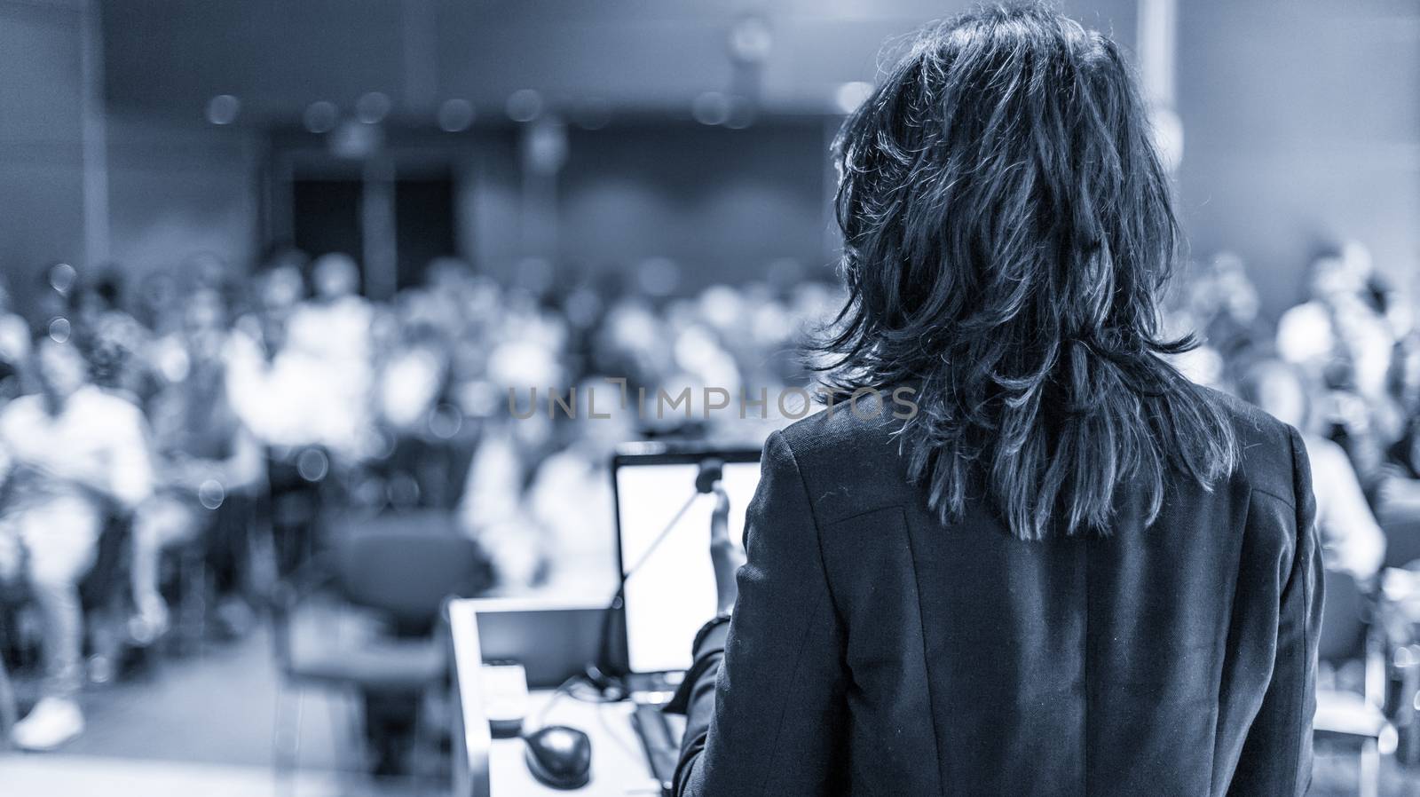 Female speaker giving a talk on corporate business conference. Unrecognizable people in audience at conference hall. Business and Entrepreneurship event. Black and white, blue toned image.