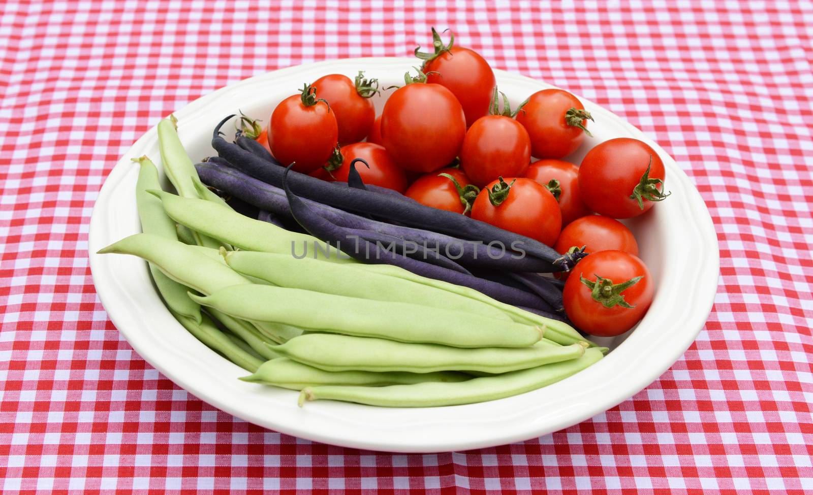 Green yin yang beans, purple French beans and juicy red cherry tomatoes in a round white dish on a red gingham tablecloth