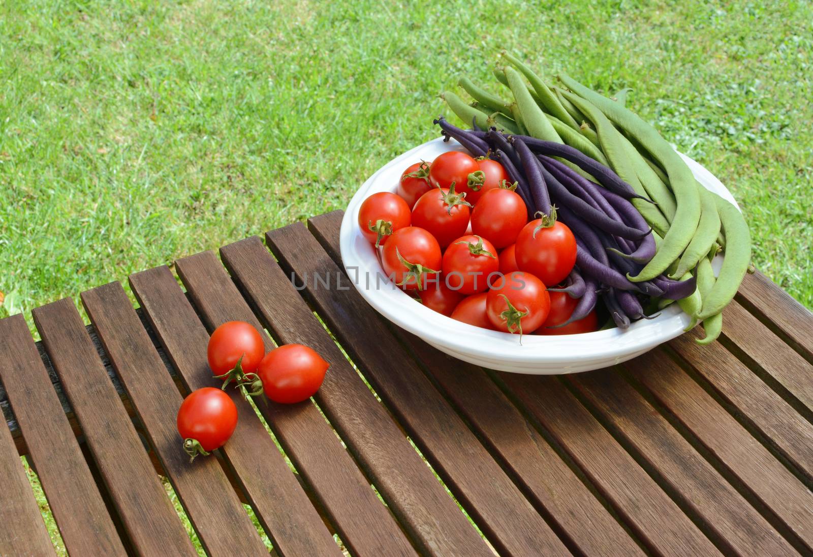 Red tomatoes with green and purple beans in a dish, some cherry tomatoes spilled on a wooden picnic table 