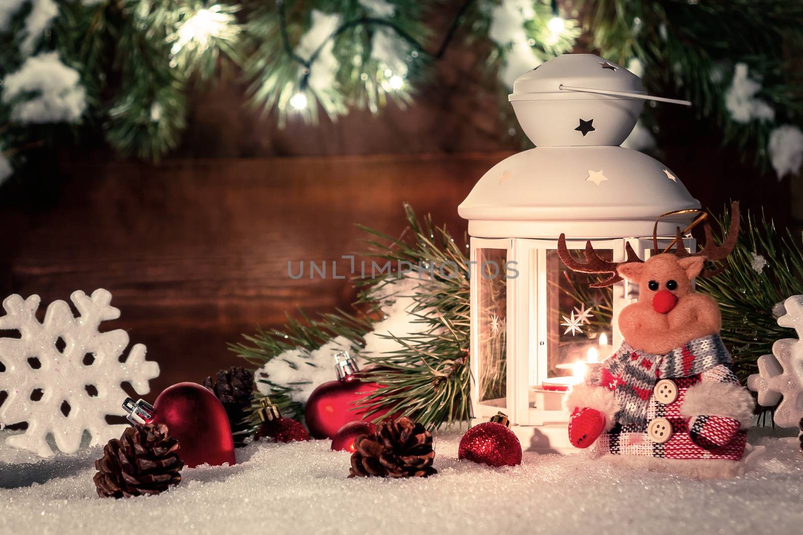 White lantern with a burning candle stands in the snow surrounded by Christmas decorations on the background of a wooden wall, Christmas tree branches and lights by galsand