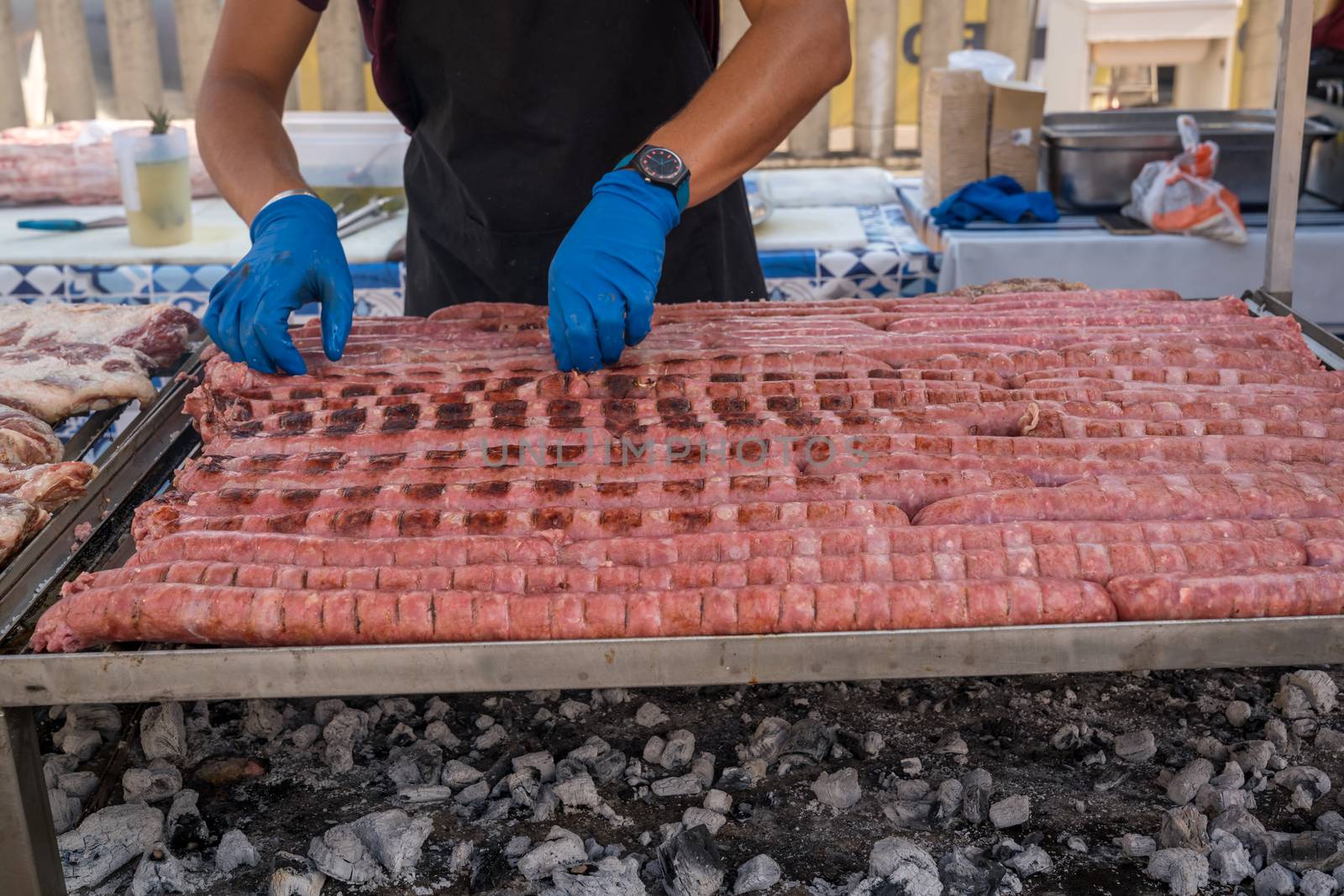 BBQ Sausage lined up on a large outdoor grill with smoke coming up from the fire below. person in black apron blue shirt gloved hand holding tongs turning meat