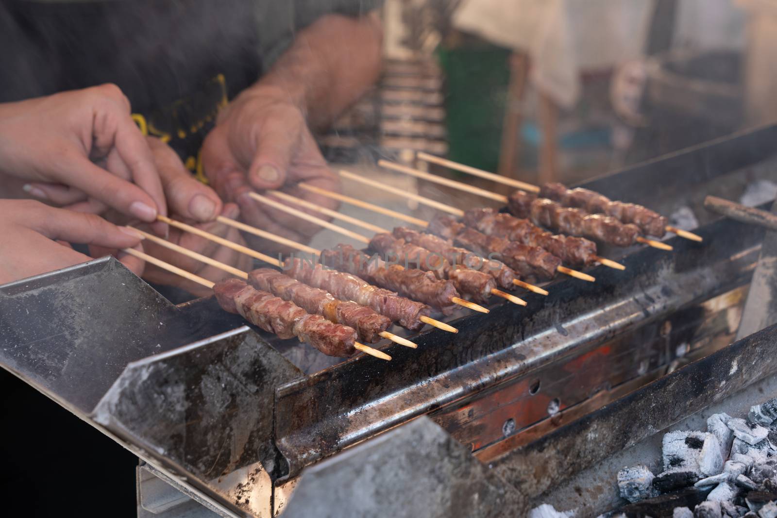 Man cooking marinated meat, Pork meat grilling on wooden skewer, outdoor grill with smoke coming up from the fire below. close up