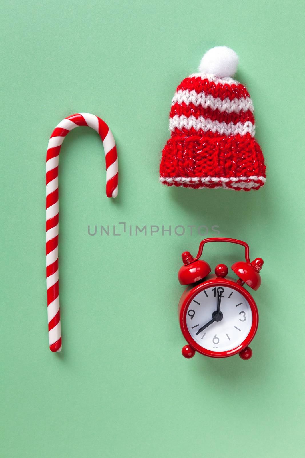 Christmas candy cane, clock and hat on pastel green backdrop. Winter minimalistic flat lay. Vertical. Top view. Still life. Waiting for holiday. Close-up, top view.