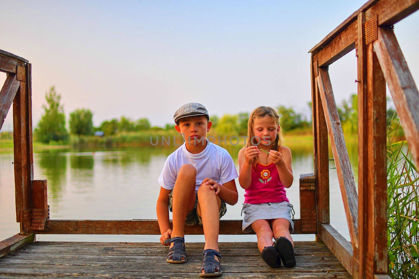 Children sitting on pier. Two children of different age - elementary age boy and preschool girl sitting on a wooden pier. Summer and childhood concept. Children on bench at the lake.