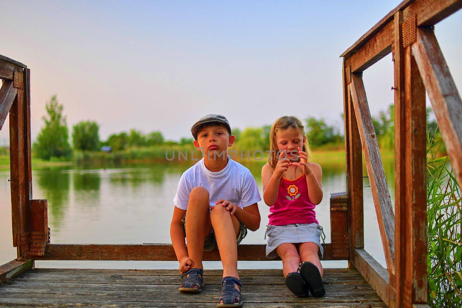 Children sitting on pier. Two children of different age - elementary age boy and preschool girl sitting on a wooden pier. Summer and childhood concept. Children on bench at the lake.