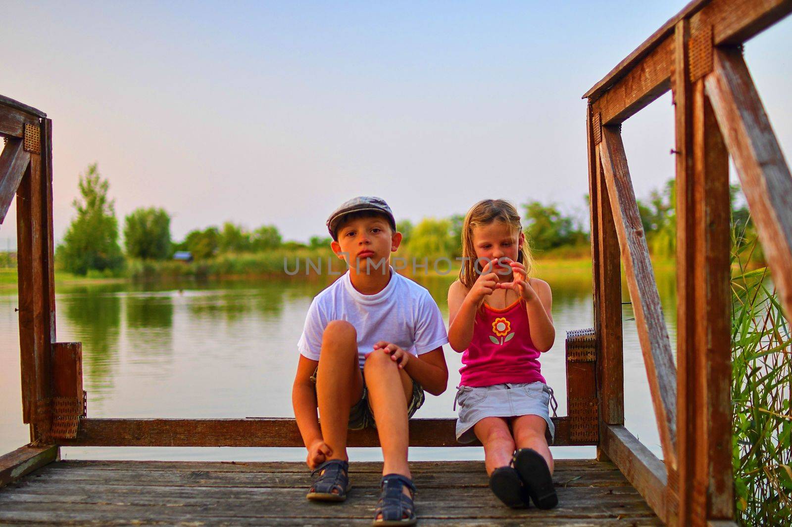 Children sitting on pier. Two children of different age - elementary age boy and preschool girl sitting on a wooden pier. Girl making heart shape. Summer and childhood concept. Children on bench at the lake.