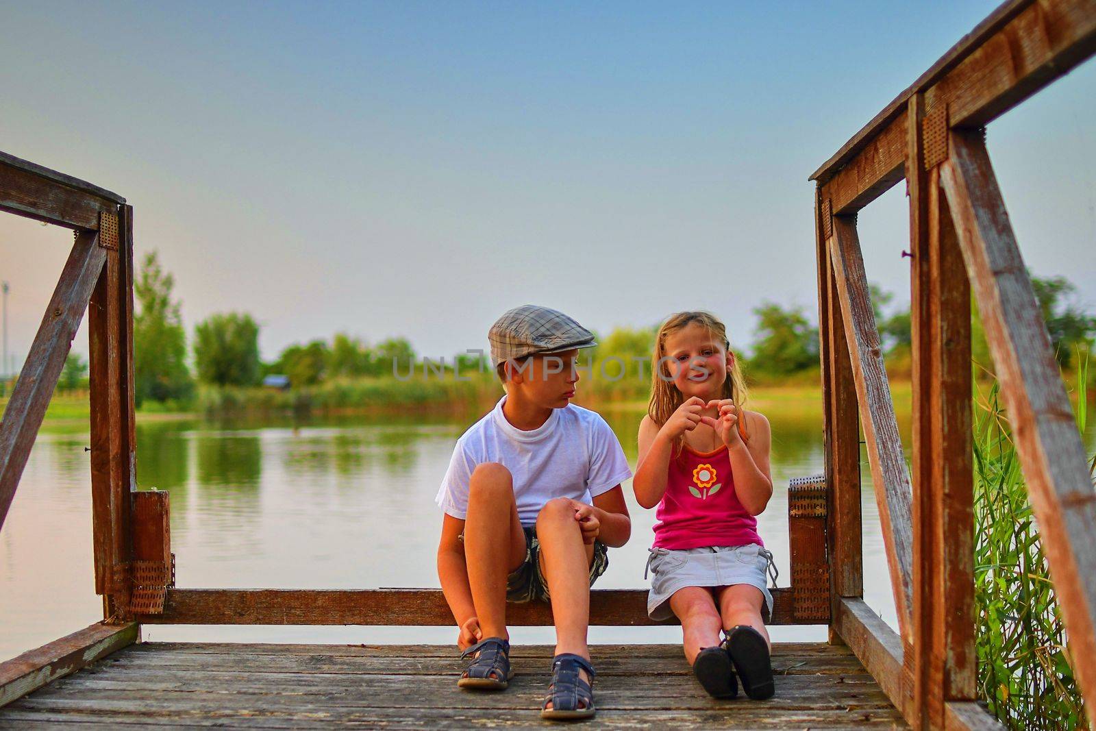 Children sitting on pier. Two children of different age - elementary age boy and preschool girl sitting on a wooden pier. Girl making heart shape. Summer and childhood concept. Children on bench at the lake.