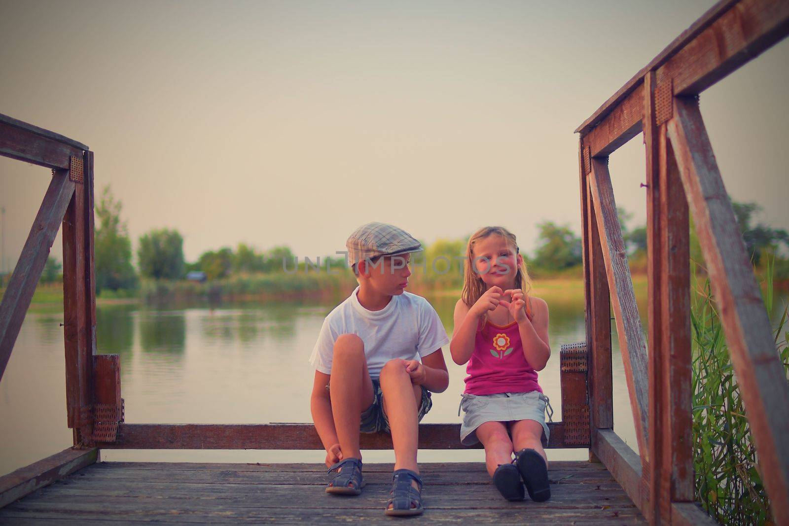 Children sitting on pier. Two children of different age - elementary age boy and preschool girl sitting on a wooden pier. Girl making heart shape. Summer and childhood concept. 