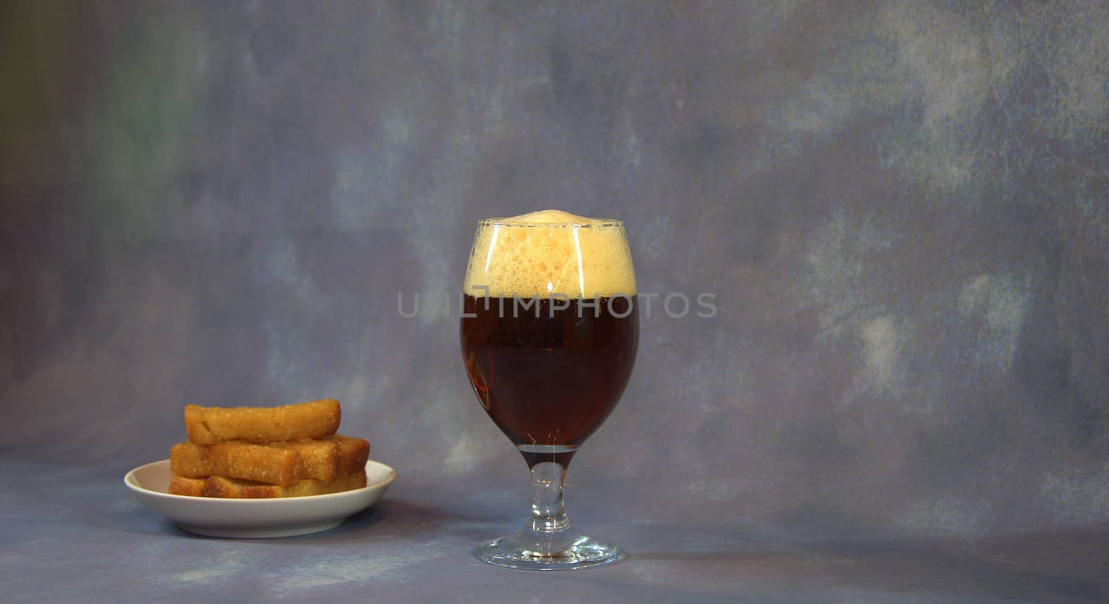 A full glass of dark beer with foam and a plate with wheat garlic croutons on a gray background. Close-up.