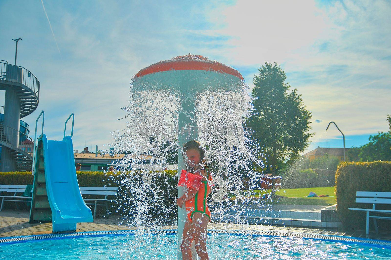 Happy little girl enjoying summer day in the swimming pool. Girl standing under sprinkler in  spray pool. Summer and happy chilhood concept. 