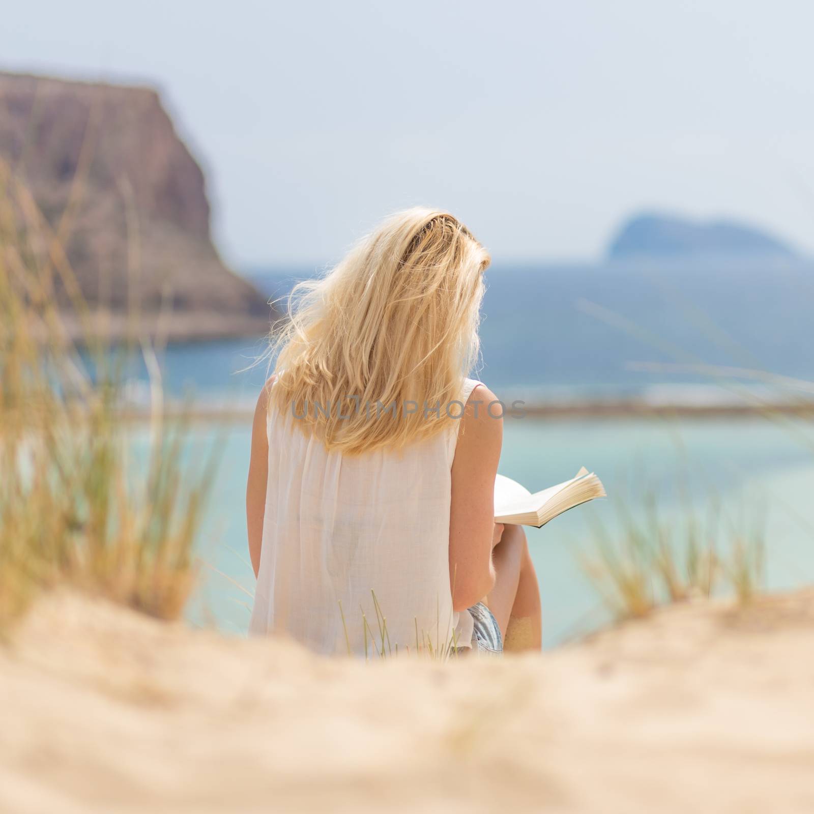 Relaxed woman enjoying sun, freedom and good book an beautiful sandy beach of Balos in Greece. Young lady reading, feeling free and relaxed. Vacations, freedom, happiness, enjoyment and well being.