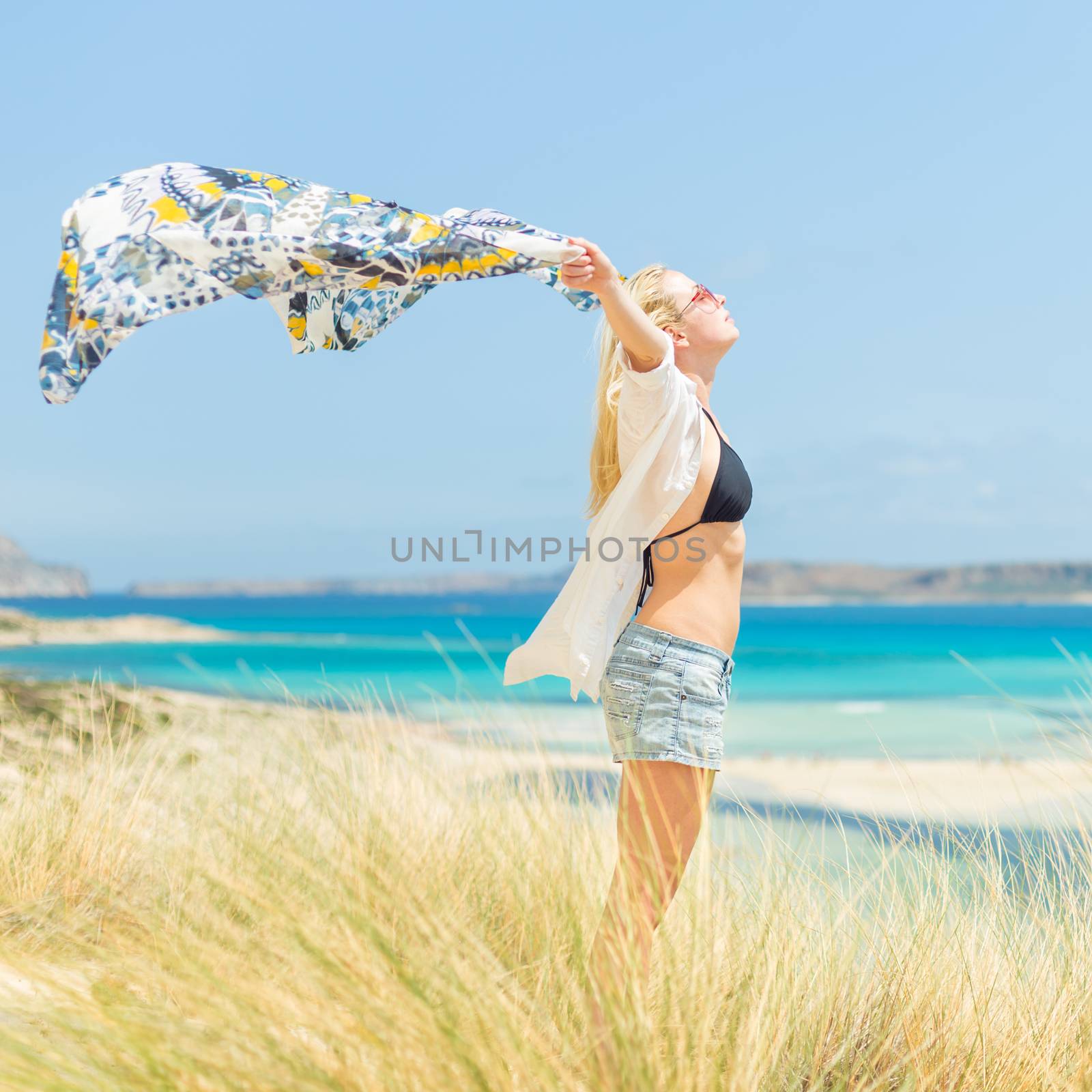 Relaxed woman, arms raised, holding colorful scarf, enjoying sun, freedom and life at beautiful beach. Young lady feeling free, relaxed and happy. Concept of vacations, freedom, joy and well being.