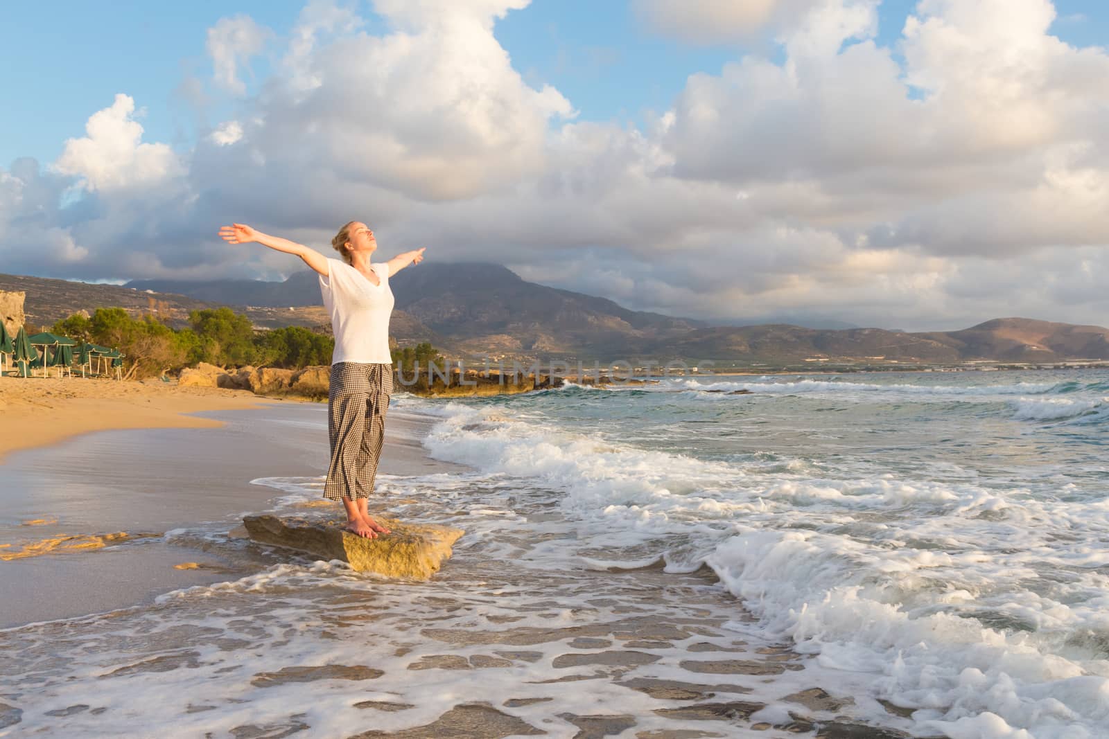 Relaxed woman, arms rised, enjoying sun, freedom and life an beautiful beach in sunset. Young lady feeling free, relaxed and happy. Concept of vacations, freedom, happiness, enjoyment and well being.