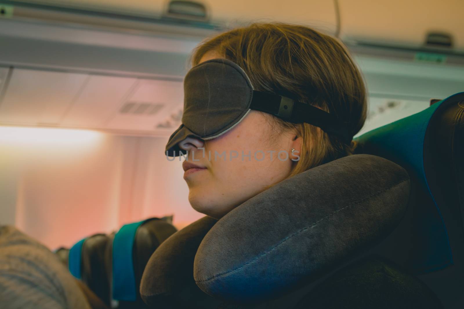 Tired caucasian blond woman with neck pillow and blindfold sleeping on a seat while traveling in a commercial airplane.