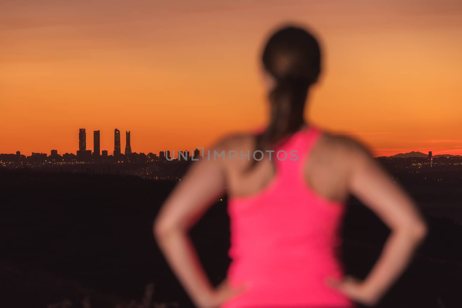 Woman on sport clothes watching sunset over city skyline. Focus is on background. by HERRAEZ