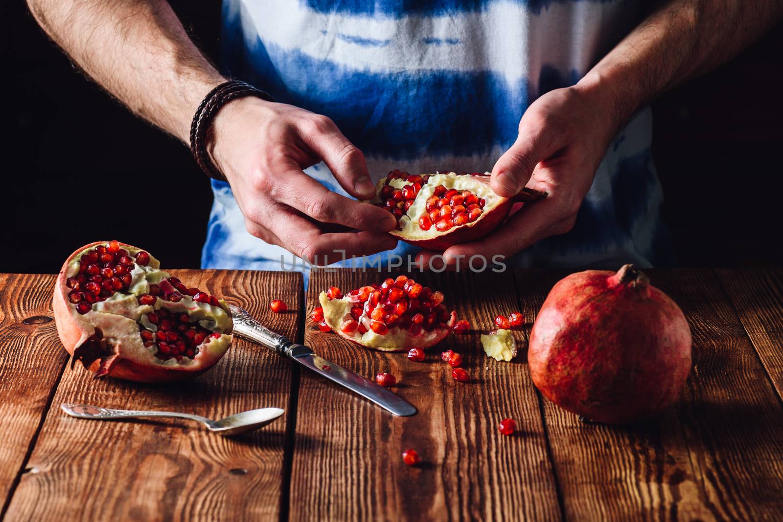 Opened Pomegranate Fruit in Hands and Other Pieces with Knife and Spoon on the Table