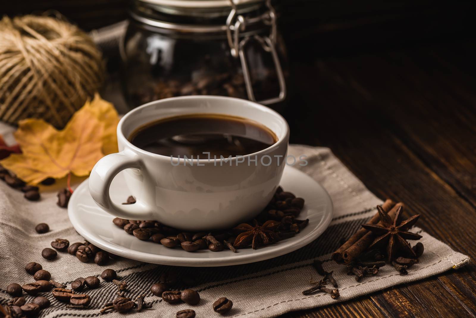 White cup of coffee with various spices, beans and yellow autumn leaves on dark wooden background