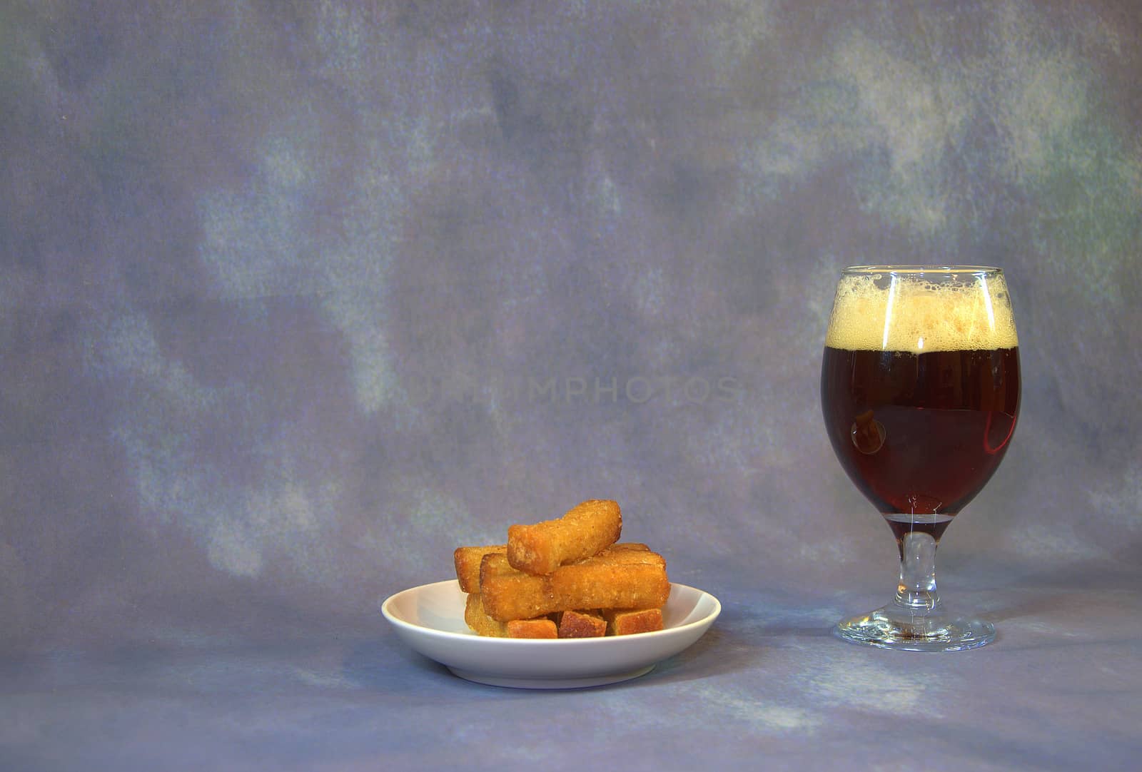 A glass of dark beer with white foam, next to a plate with wheat croutons. Close-up.