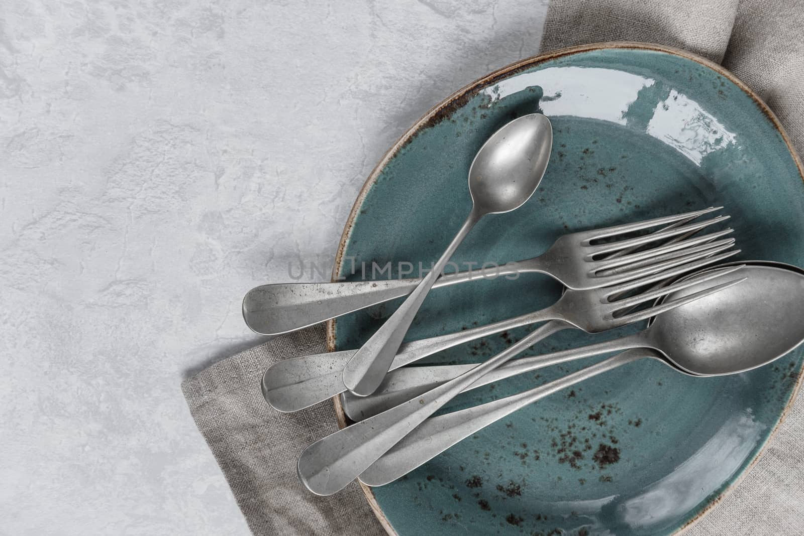 Various silverware on a blue ceramic plate and gray flax napkin are on the background of gray concrete surface, with copy-space